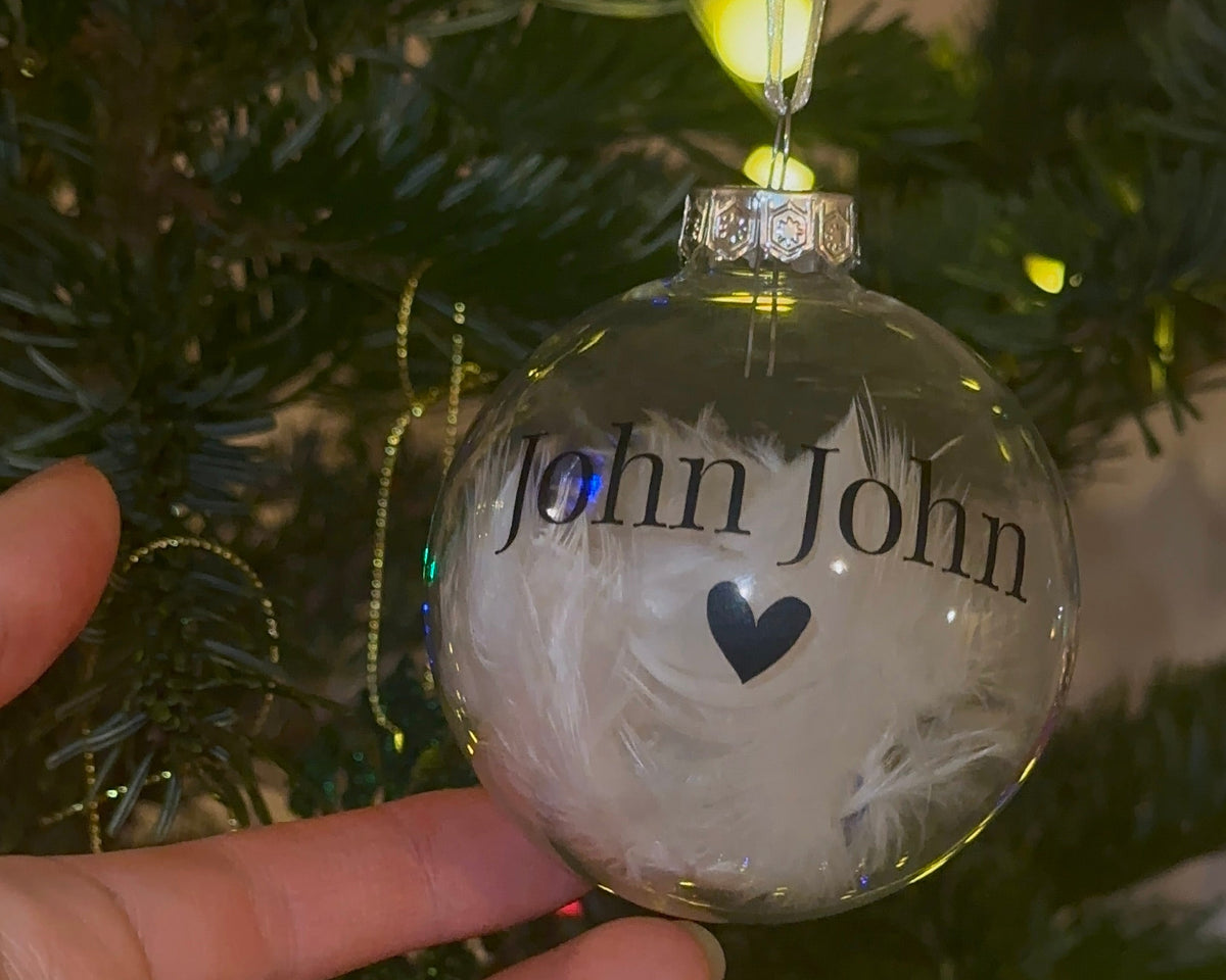 A clear round Christmas ornament with white feathers and the text 'John John' and a small black heart, hanging on a decorated Christmas tree while a hand holds it.