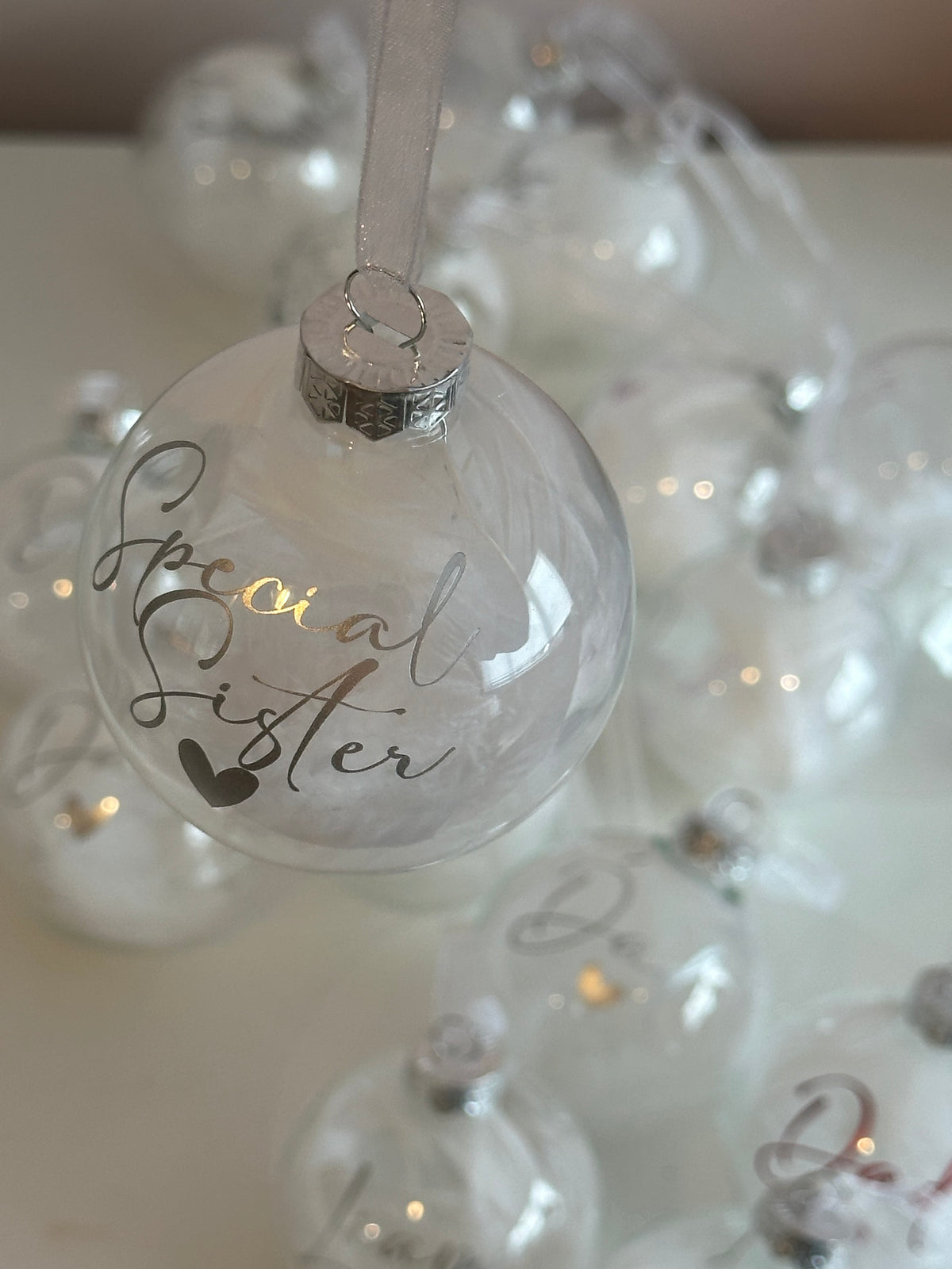 A clear glass Christmas ornament hanging by a white ribbon, printed with the words 'Special Sister' in black script and a small heart, with warm white fairy lights glowing inside. Other similar ornaments are softly blurred in the background.
