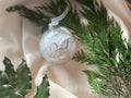 A clear glass Christmas ornament with white feathered detailing and the word 'Dad' written in elegant script, styled on cream satin fabric with green pine branches and holly leaves.