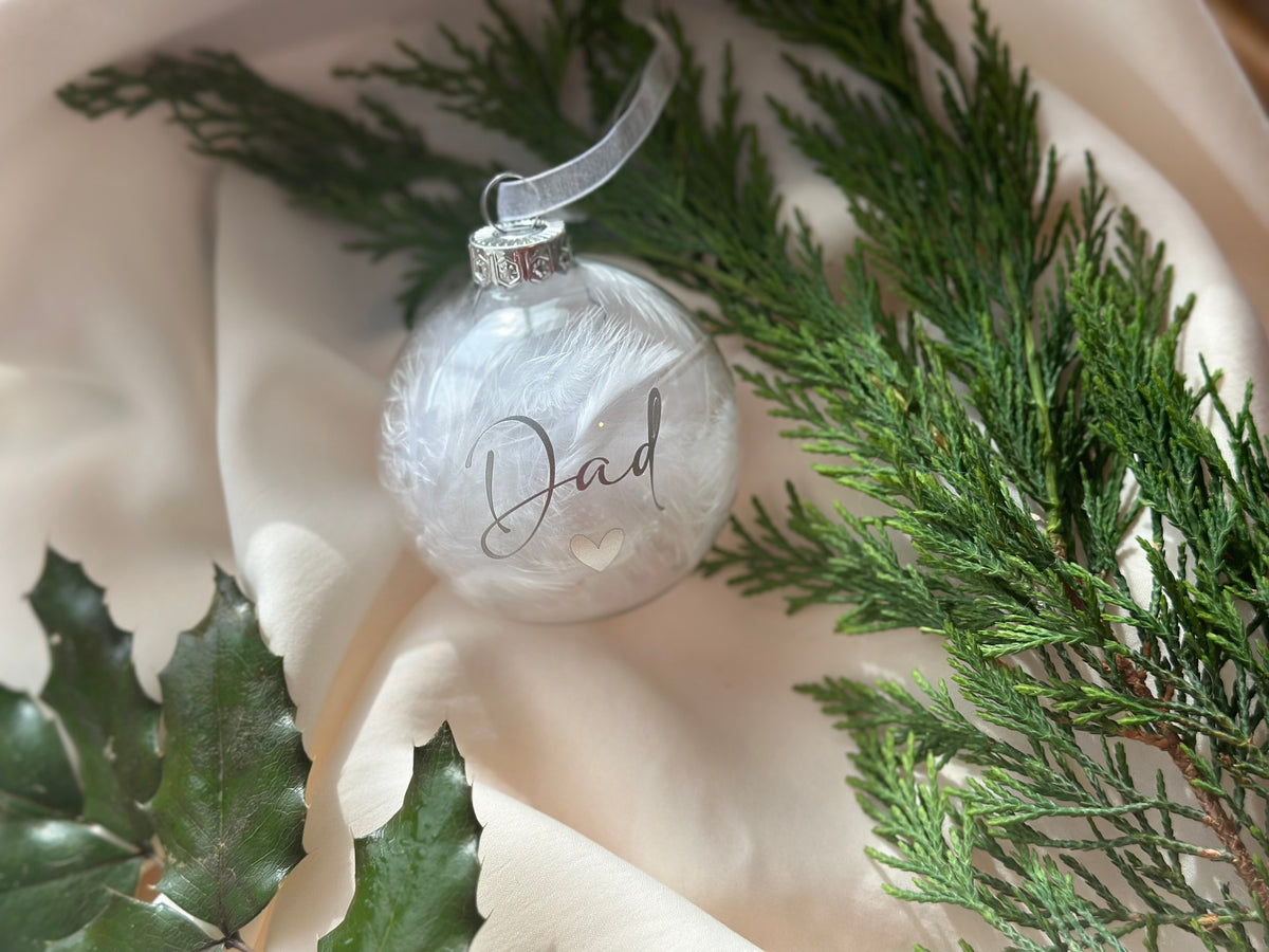 A clear glass Christmas ornament with white feathered detailing and the word 'Dad' written in elegant script, styled on cream satin fabric with green pine branches and holly leaves.