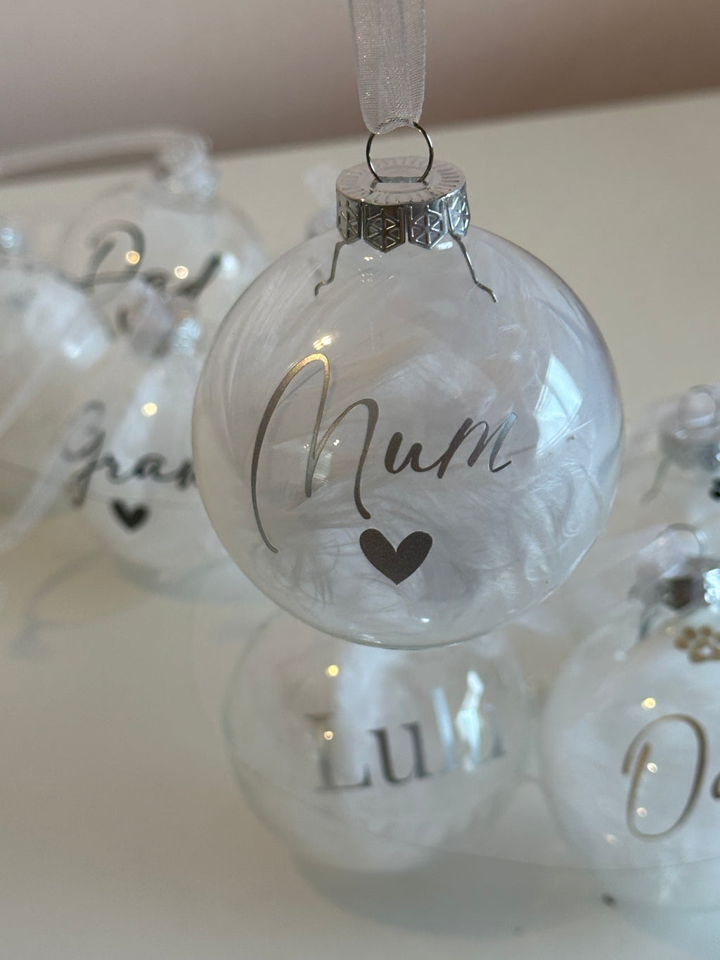 A clear round Christmas ornament with silver lettering reading 'Mum' and a small heart, hanging on a white surface with other similar ornaments blurred in the background.