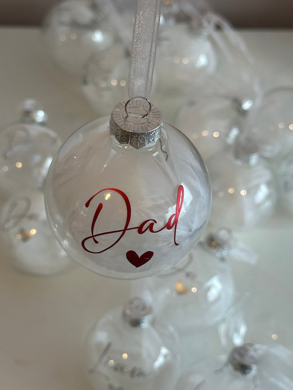A clear glass Christmas ornament hanging by a white ribbon, printed with the word 'Dad' in red cursive and a small red heart. Other similar clear ornaments are softly blurred in the background.