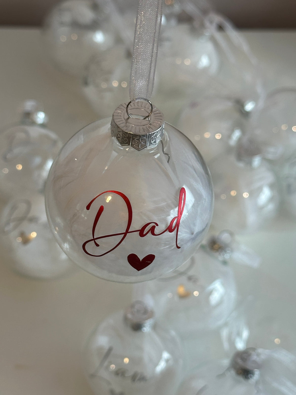 A clear glass Christmas ornament hanging by a white ribbon, printed with the word 'Dad' in red cursive and a small red heart. Other similar clear ornaments are softly blurred in the background.