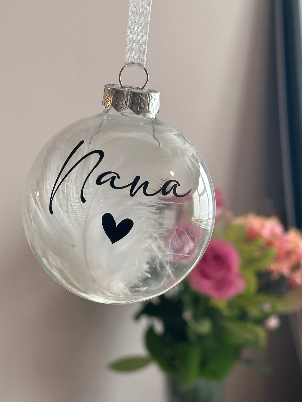 A clear glass Christmas ornament hanging from a white ribbon, featuring the word 'Nana' in black script and a small black heart, with white feathers inside. A blurred bouquet of pink flowers is visible in the background.