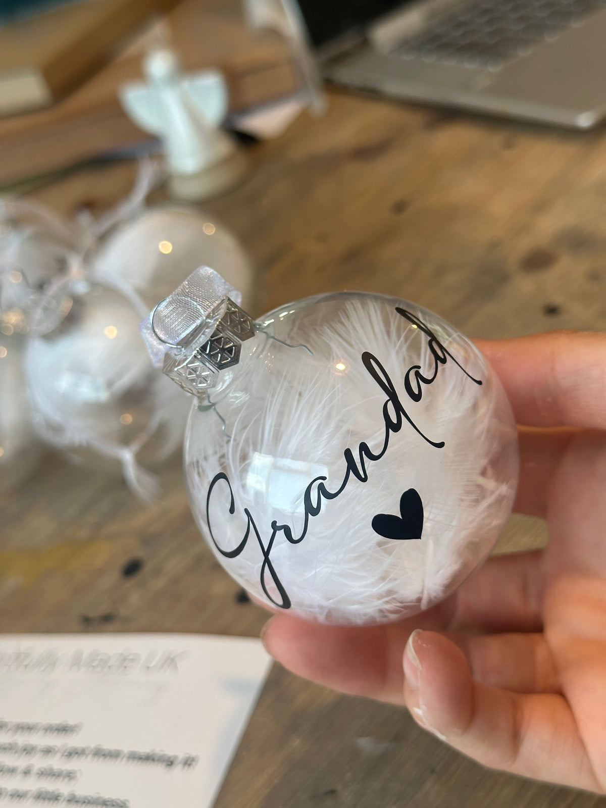 A hand holding a clear glass Christmas ornament filled with white feathers and personalized with the name 'Grandad' in black script and a small black heart. Other similar ornaments are blurred in the background on a wooden table.