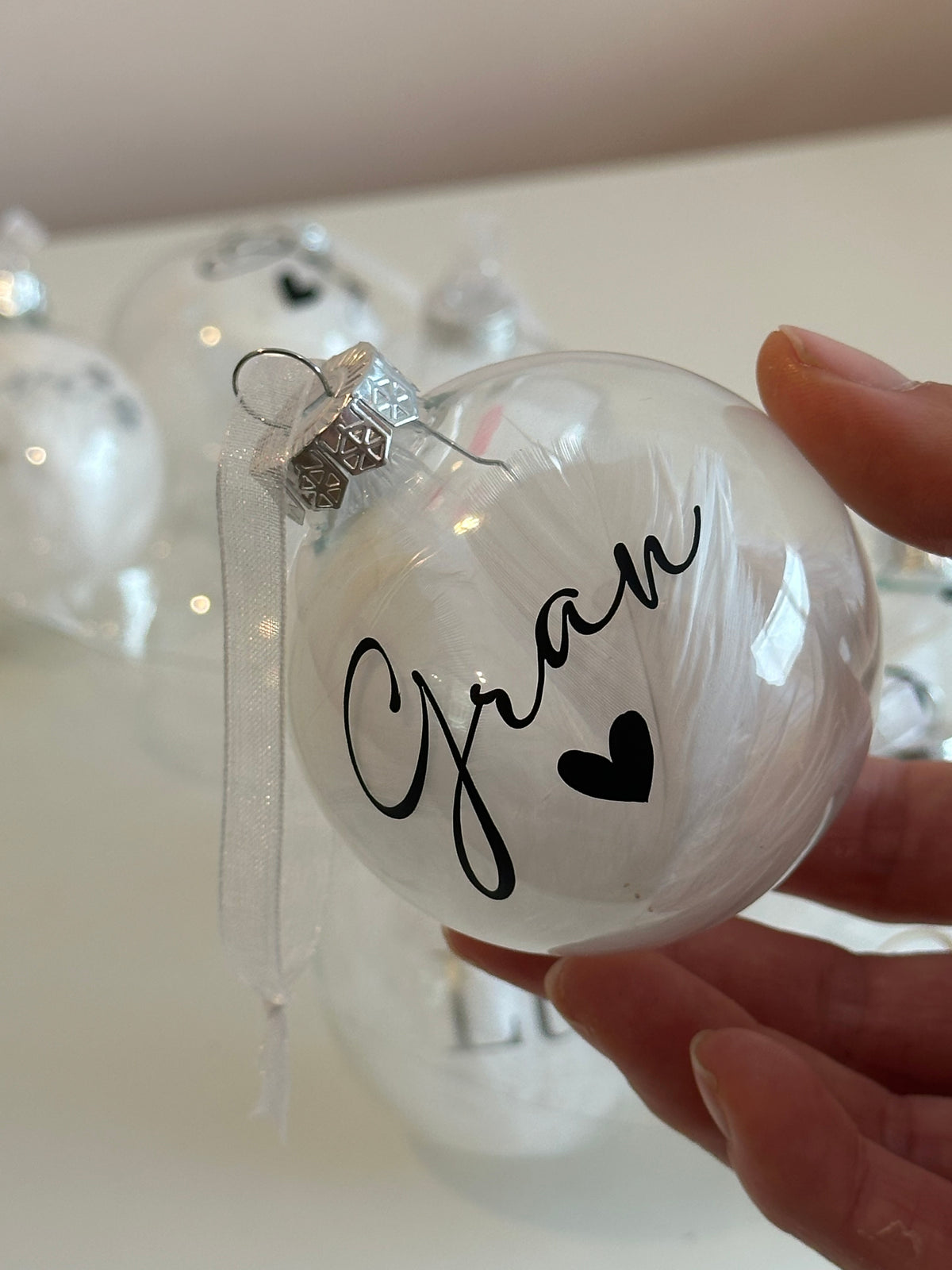 A hand holding a clear glass Christmas ornament with the name 'Gran' written in black script and a small black heart, with a silver cap and white ribbon.