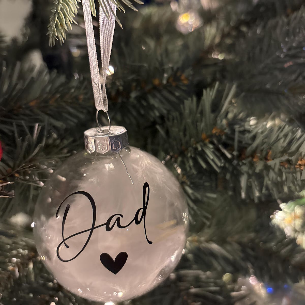 A clear round Christmas ornament hanging on a green Christmas tree, printed with the word 'Dad' in black script and a small black heart below it.