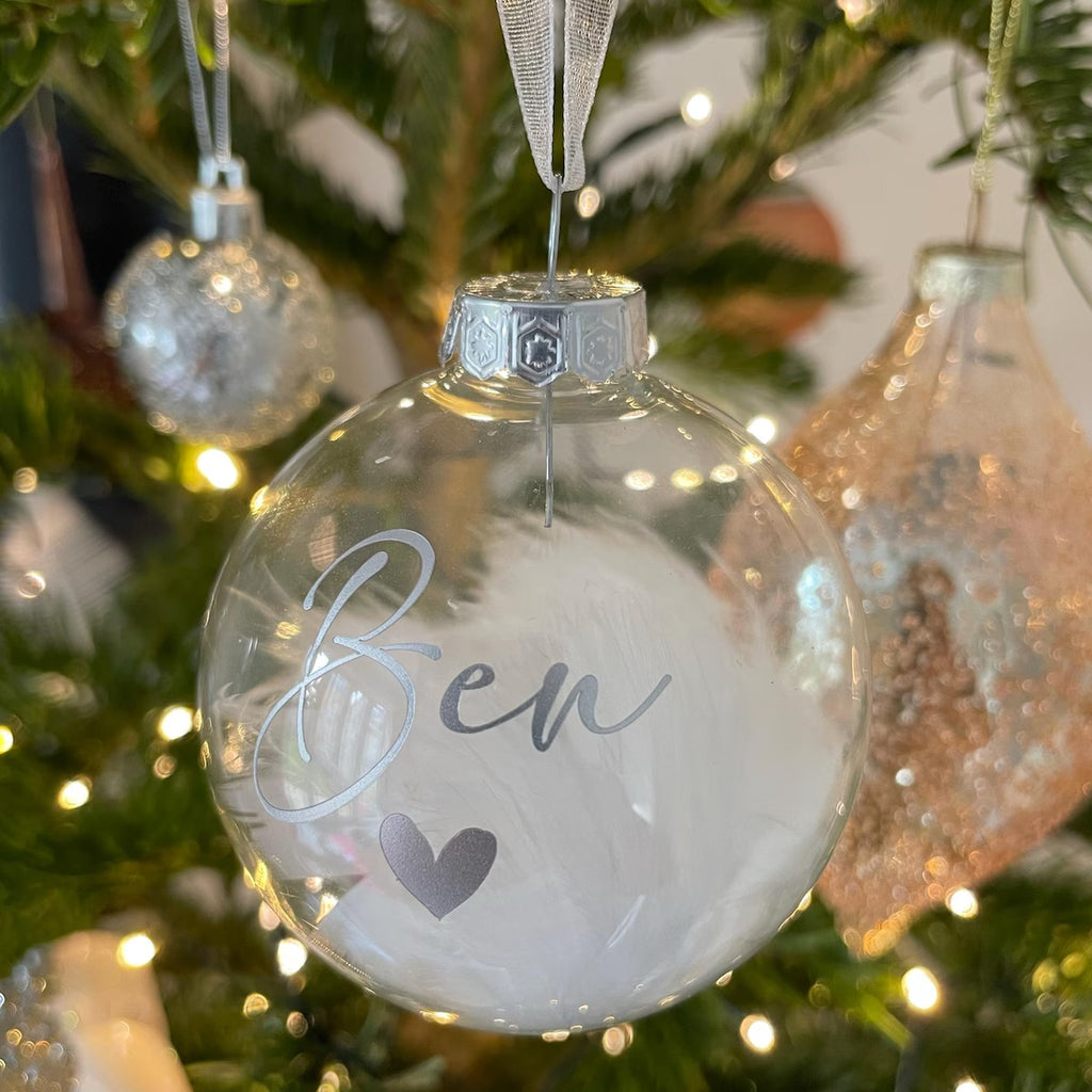 A clear round Christmas ornament with a silver cap, hanging on a decorated tree. The ornament is printed with the name 'Ben' in silver cursive and a small gray heart, with white faux fur inside.