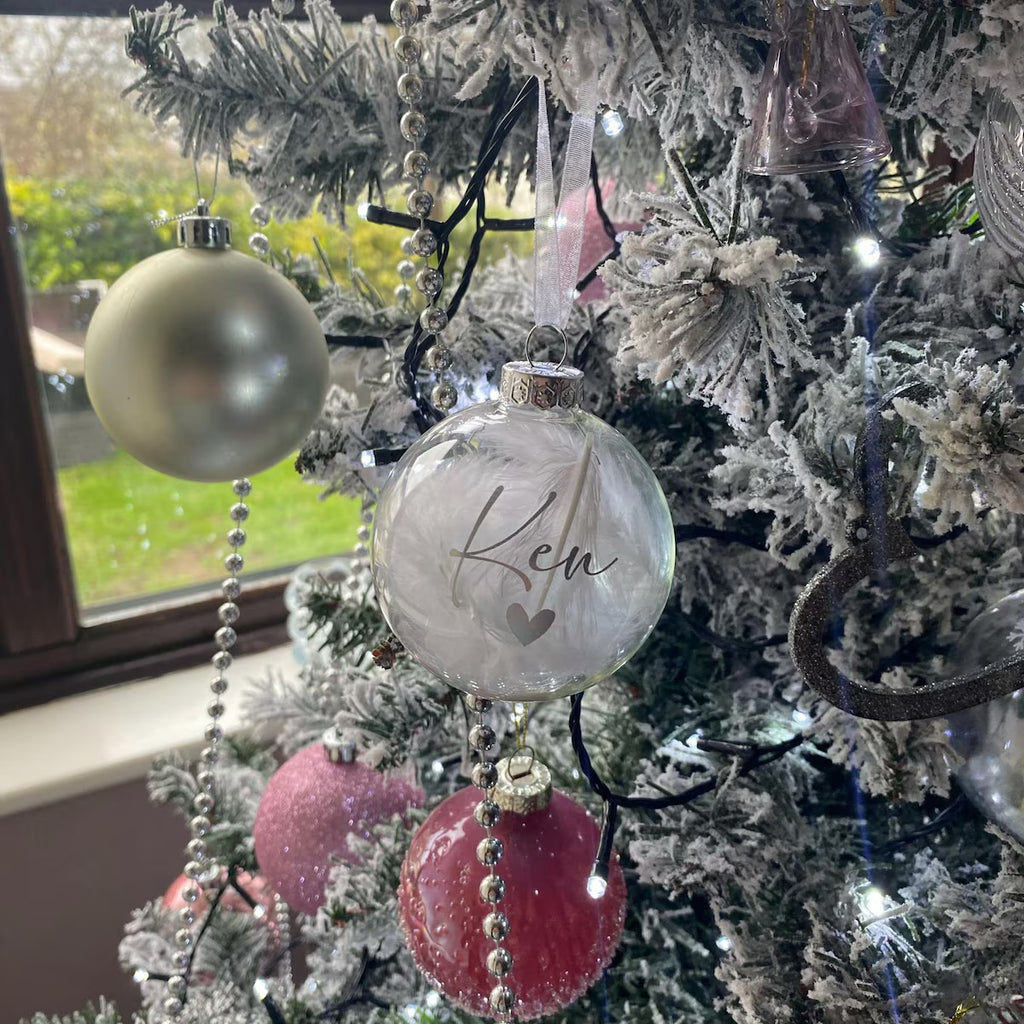 A frosted glass Christmas ornament with the name 'Ken' and a small heart printed on it, hanging on a snow-dusted Christmas tree next to a shiny silver ornament.