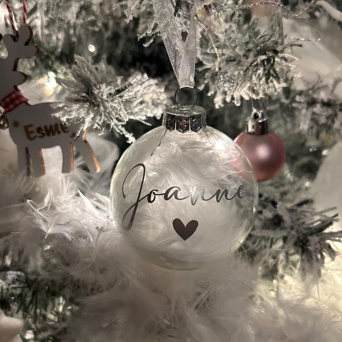 A clear round Christmas ornament hanging on a frosted white tree, printed with the name 'Joanna' in black script and a small dark heart below it. A pink ornament hangs nearby, and part of a wooden reindeer decoration is visible at left.