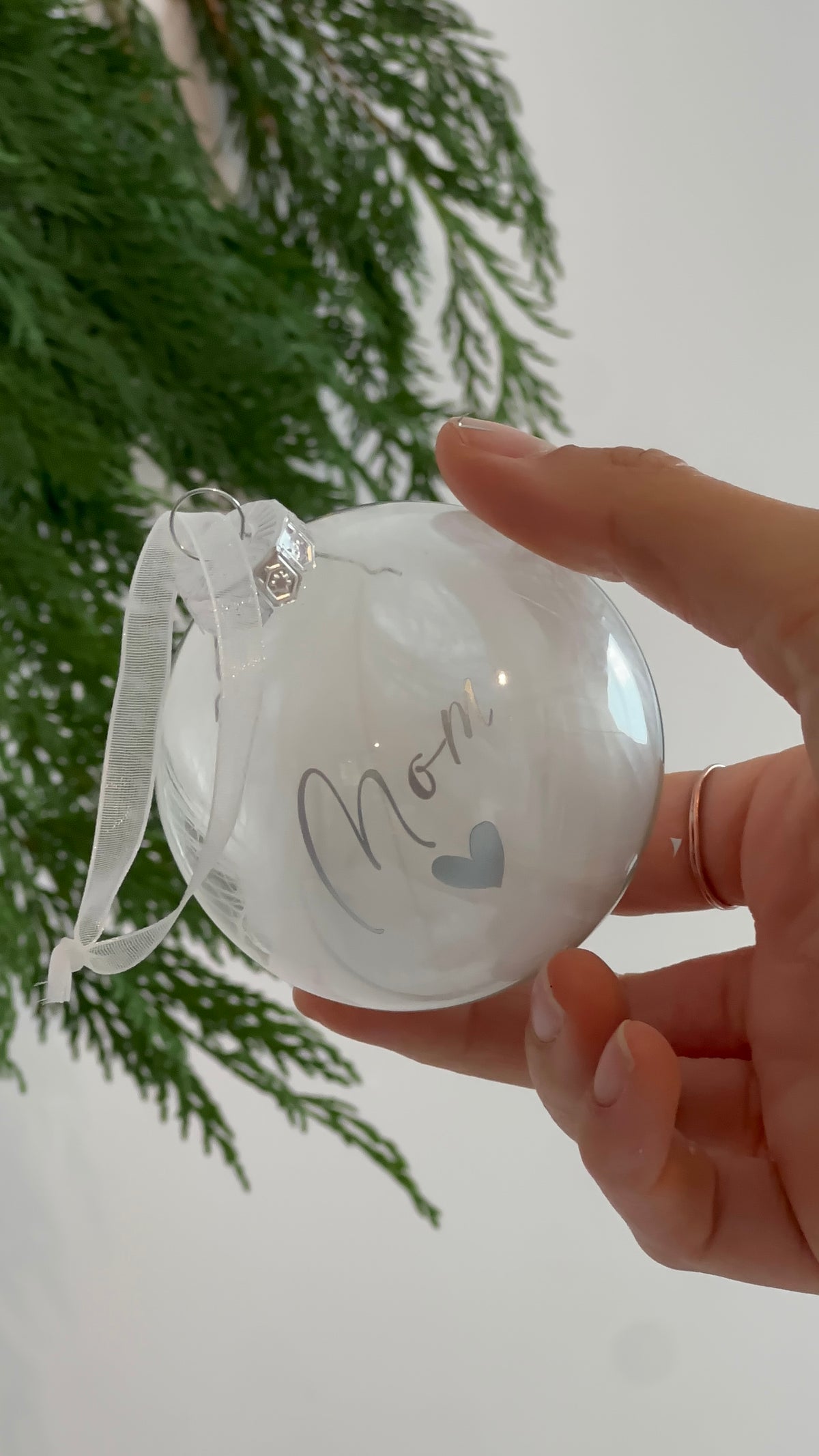 A hand holds a clear round Christmas ornament with a white ribbon hanger and the word 'Mom' written in gray script with a small gray heart. Evergreen branches hang in the background against a light wall.